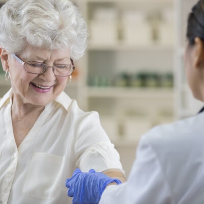 A smiling senior pharmacy customer looks down at her upper arm as a gloved pharmacist places a band-aid over her shingles vaccine shot site.