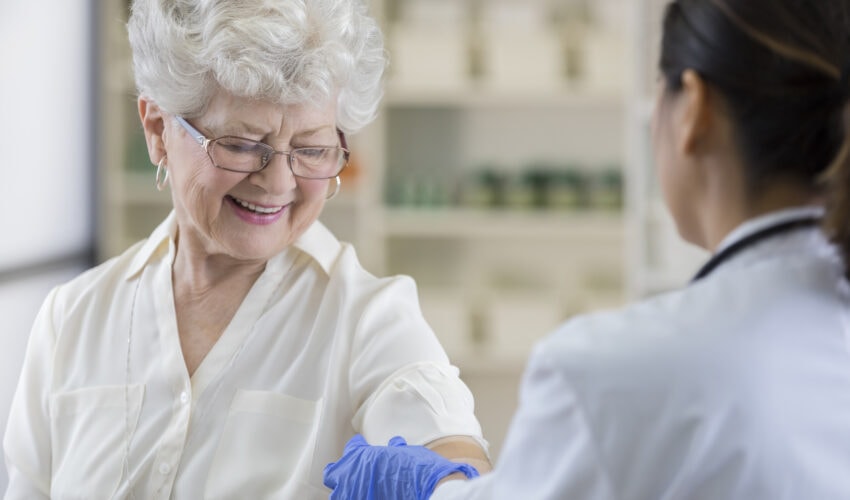 A smiling senior pharmacy customer looks down at her upper arm as a gloved pharmacist places a band-aid over her shingles vaccine shot site.